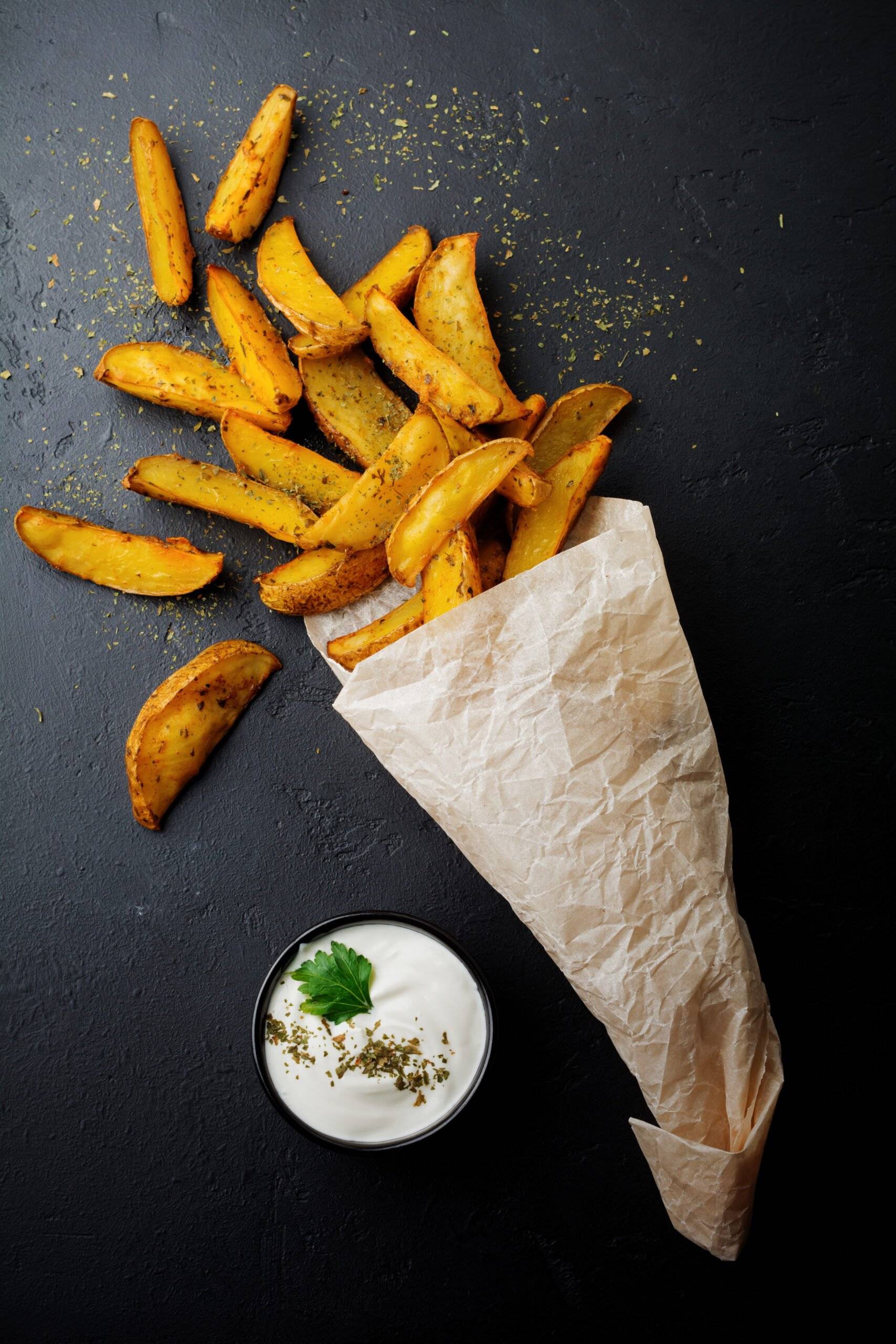 baked potatoes with greens and sauce in paper bag, scattered on dark concrete or ceramic background. selective focus. view from above. place for text